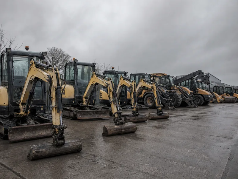 Mini excavators, telehandlers and compaction rollers lined up on hardstanding in a plant hire yard under a grey sky.